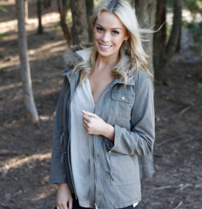 Blonde woman in an olive green, hooded parka poses outdoors in a wooded area, smiling at the camera.