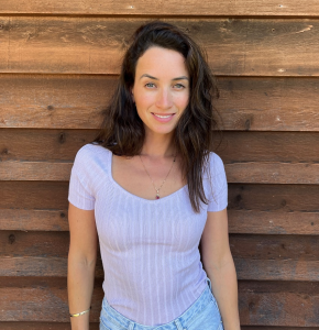 Smiling woman in a lilac top and jeans standing in front of a wooden wall, portrait, outdoor, medium shot.