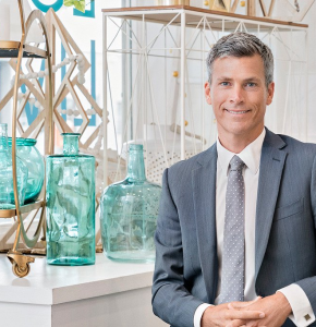 Portrait of a smiling Caucasian adult male in a suit in an indoor setting with decorative glass bottles on a white shelf.