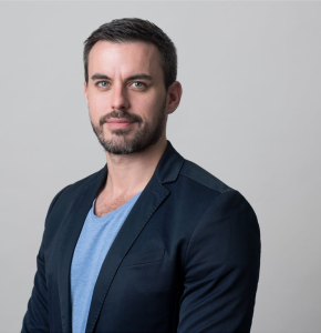 Headshot of a confident adult male with dark hair, wearing a blue blazer and light blue t-shirt against a neutral background.