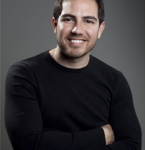 Headshot of an adult male with arms crossed, wearing a black long-sleeve shirt against a grey background in a photo studio.