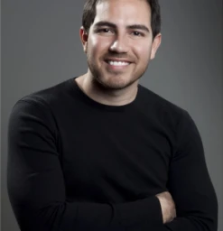 Portrait of a man with short dark hair and a beard wearing a black long-sleeve shirt against a dark grey background.
