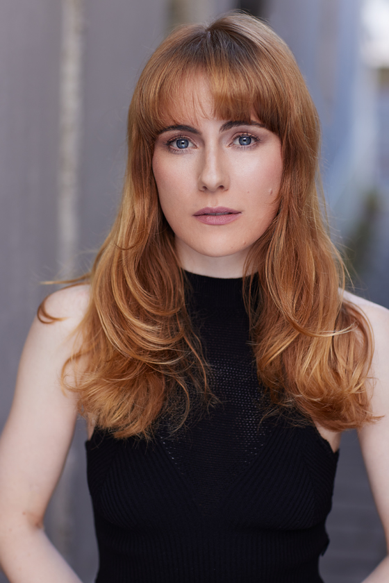 Portrait of a woman with long auburn hair and a black top against a blurred background, looking directly at the camera.