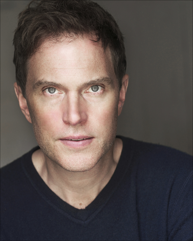 Portrait of a fair-skinned adult male with short brown hair and blue eyes, wearing a dark blue v-neck sweater against a gray background.