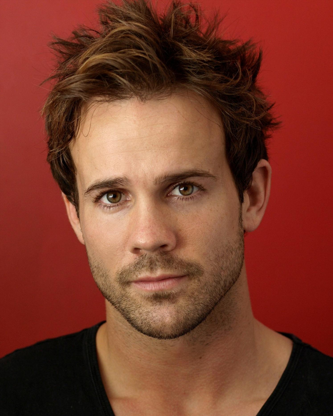 Close-up studio portrait of a handsome adult male with light brown hair, wearing a black v-neck shirt, against a red backdrop.
