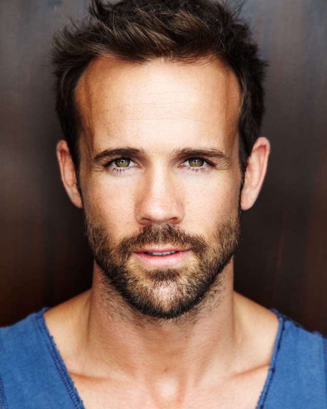 Headshot of a light-skinned adult man with short brown hair and beard, wearing a blue shirt, against a neutral background.