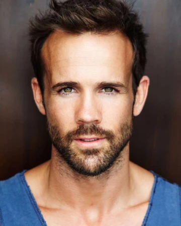 Close-up portrait of a man with short brown hair, beard, and blue shirt against a dark textured background