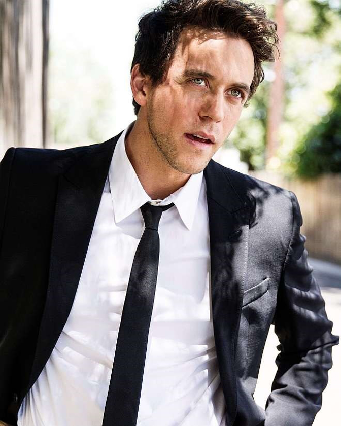 Portrait of a man wearing a black suit with a white shirt and black tie, posing outdoors with natural lighting and blurred background.