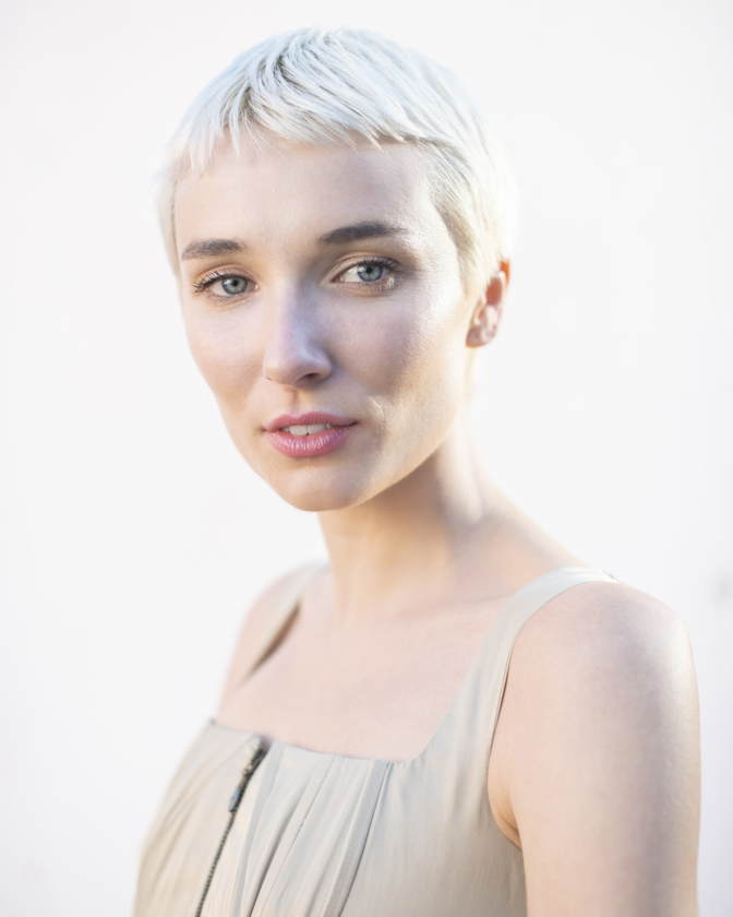 Close-up portrait of a woman with short, white-blonde hair and blue eyes, wearing a light gray dress against a bright, neutral background.