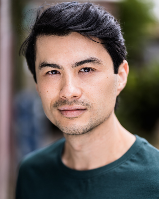 Portrait of a young adult male with dark hair wearing a green shirt, looking directly at the camera against a blurred background.