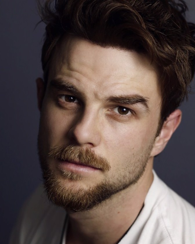 Headshot of a handsome adult male with brown hair, a beard, and a white shirt, set against a dark gray background in a portrait orientation.