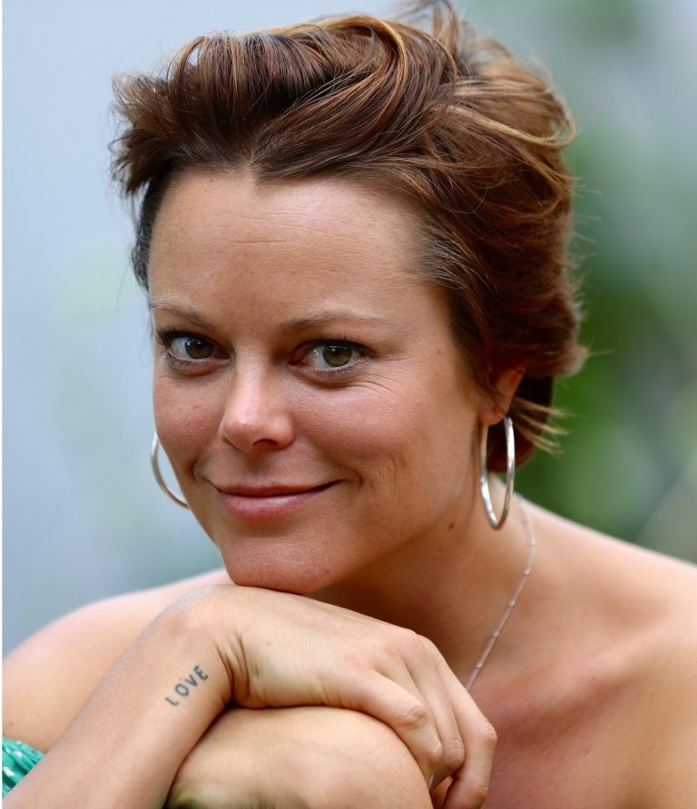 Close-up portrait of an adult woman with short hair, wearing hoop earrings, fair skin, and a tattoo on her left arm outdoors.
