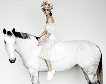 Fashion shot of a woman in a white dress and flower crown riding a white horse in a bright studio setting, showcasing elegance and style.