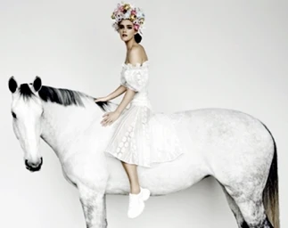 A woman wearing a floral crown and white dress sitting on a white horse against a plain seamless background.
