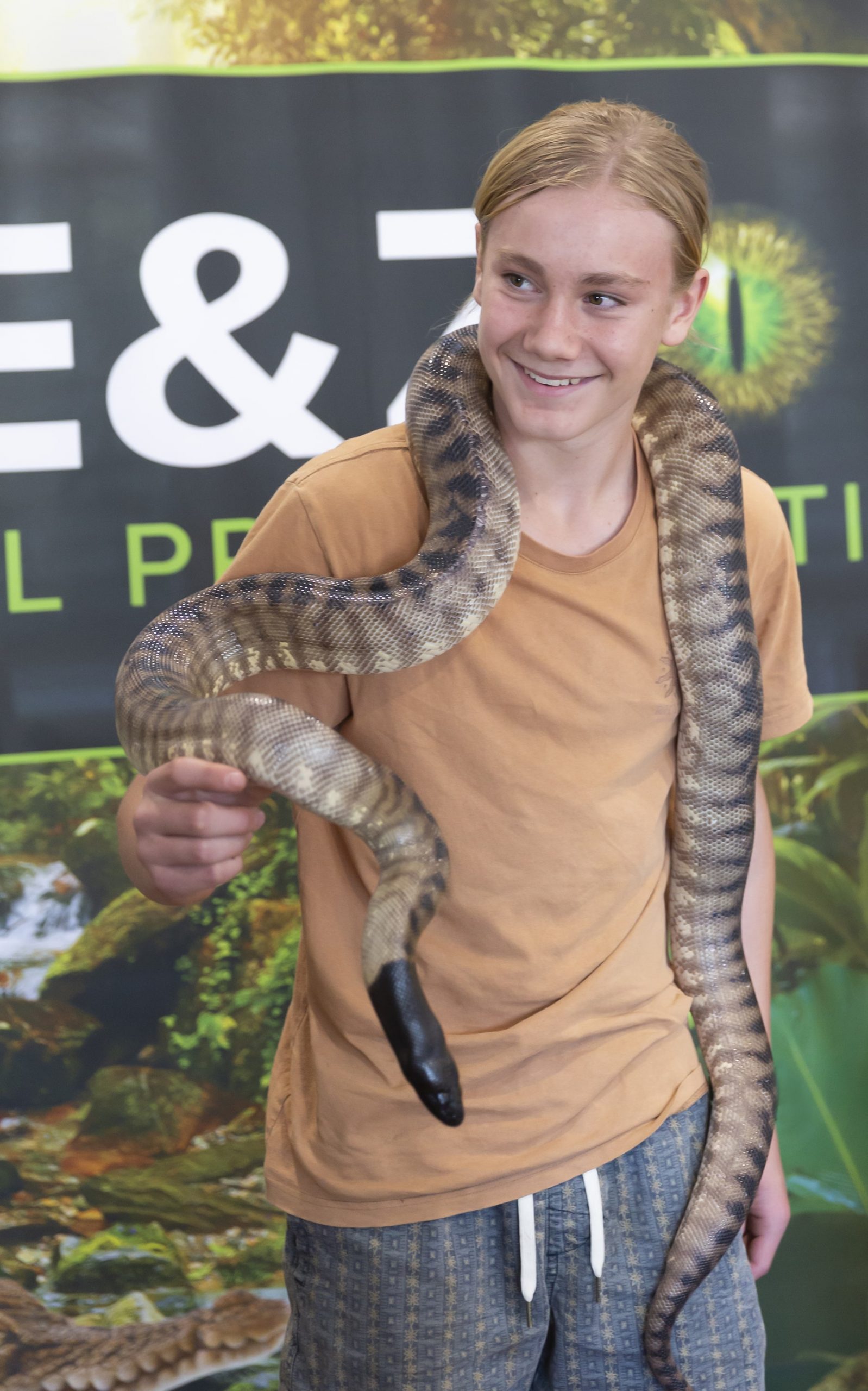 A smiling child poses with a python draped around their shoulders in an indoor setting with a nature scene in the backdrop.