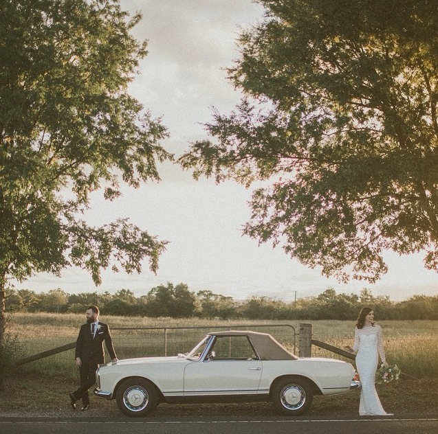 Bride and groom pose beside vintage convertible car on rural road with trees and field at golden hour on their wedding day.