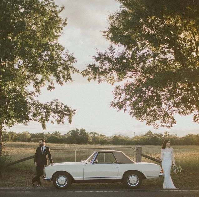 Bride and groom pose beside vintage convertible car on rural road with trees and field at golden hour on their wedding day.
