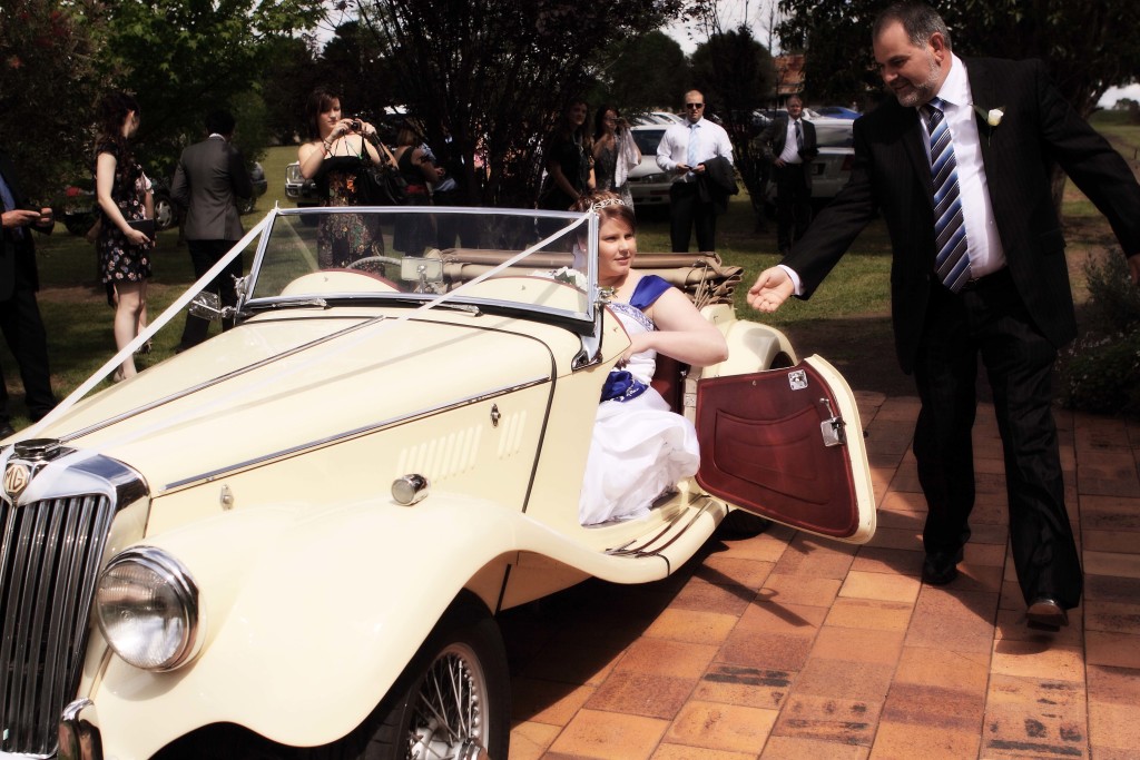 Bride and man near vintage car with wedding decorations, surrounded by guests outside on a sunny day, shot with a medium focal length.