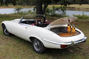White vintage Jaguar E-Type convertible with the trunk open displaying two whiskey bottles, set in a natural outdoor landscape.