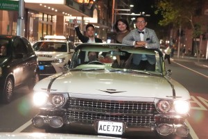 Three adults in a vintage Cadillac convertible at night on a city street, with bright headlights and warm city lights in the background.