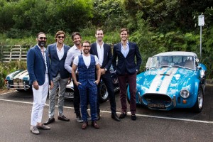 Group of six men in suits standing with classic cars outdoors, sunny day, professional attire, automotive event, daytime scene.