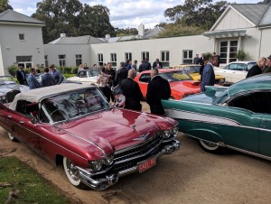 Classic cars on display in front of a building, with a crowd of people in suits visible in the background, under an overcast sky.