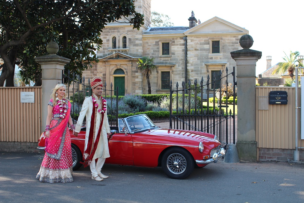 An Indian couple in traditional wedding attire stand next to a red convertible in front of a stone building on a sunny day.