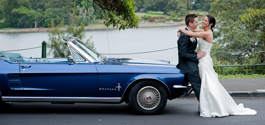 A smiling couple in wedding attire embraces next to a classic blue Mustang convertible with a waterfront and tree-lined backdrop.