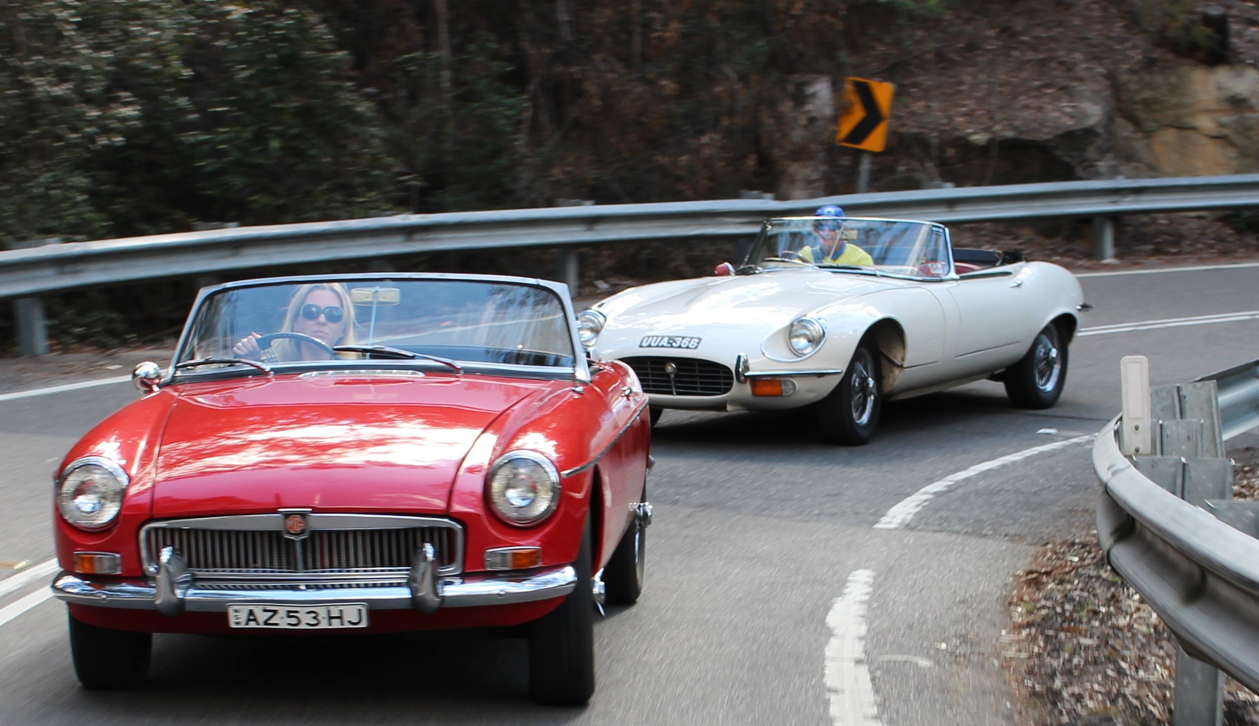 Two classic convertible cars, one red and one white, drive on a winding road surrounded by trees and a guardrail, on a sunny day.