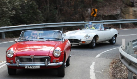 Vintage red sports car driving on a winding mountain road followed by a white convertible