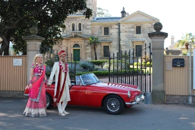 Bride in a pink lehenga and groom in a cream sherwani standing beside a vintage red convertible car in Sydney.