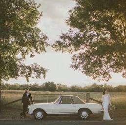 Bride and groom posing by a vintage white convertible car in an open field framed by trees