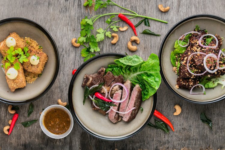 Overhead shot of three plates of prepared foods: fried appetizers, sliced steak, and dark meat with cashew garnish and greens.