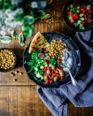 Overhead shot of a blue bowl filled with chickpeas, olives, figs, greens, and pita bread on a wooden table with a grey cloth.