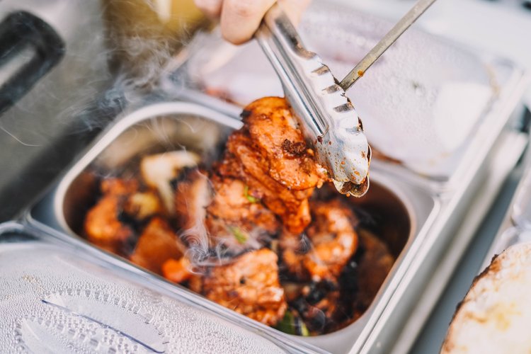 Close-up of steaming, seasoned chicken being lifted from a stainless-steel food pan with metal tongs, likely at a catered event.