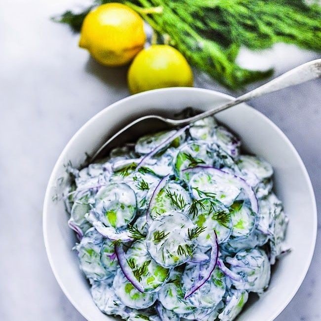 Overhead shot of a white bowl filled with creamy cucumber salad with dill and red onion, lemons and fresh dill in background.