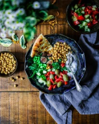 Overhead food shot of a colorful grain bowl with chickpeas, figs, olives, and greens served on a rustic dark wood table.