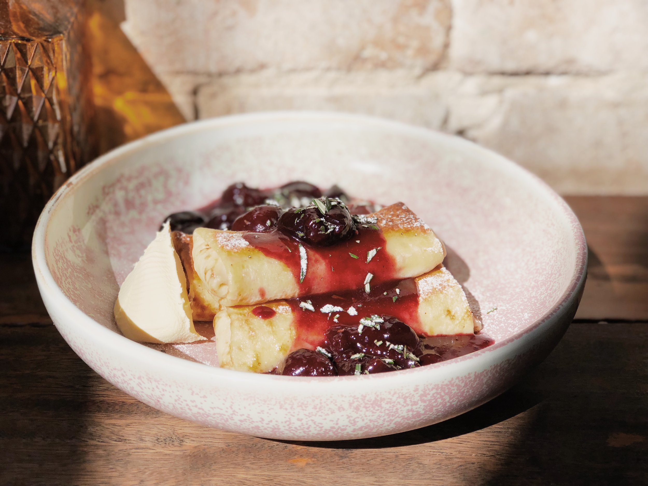 Overhead shot of French toast slices topped with cherry sauce in a speckled ceramic bowl, set on a rustic wooden table, natural sunlight.
