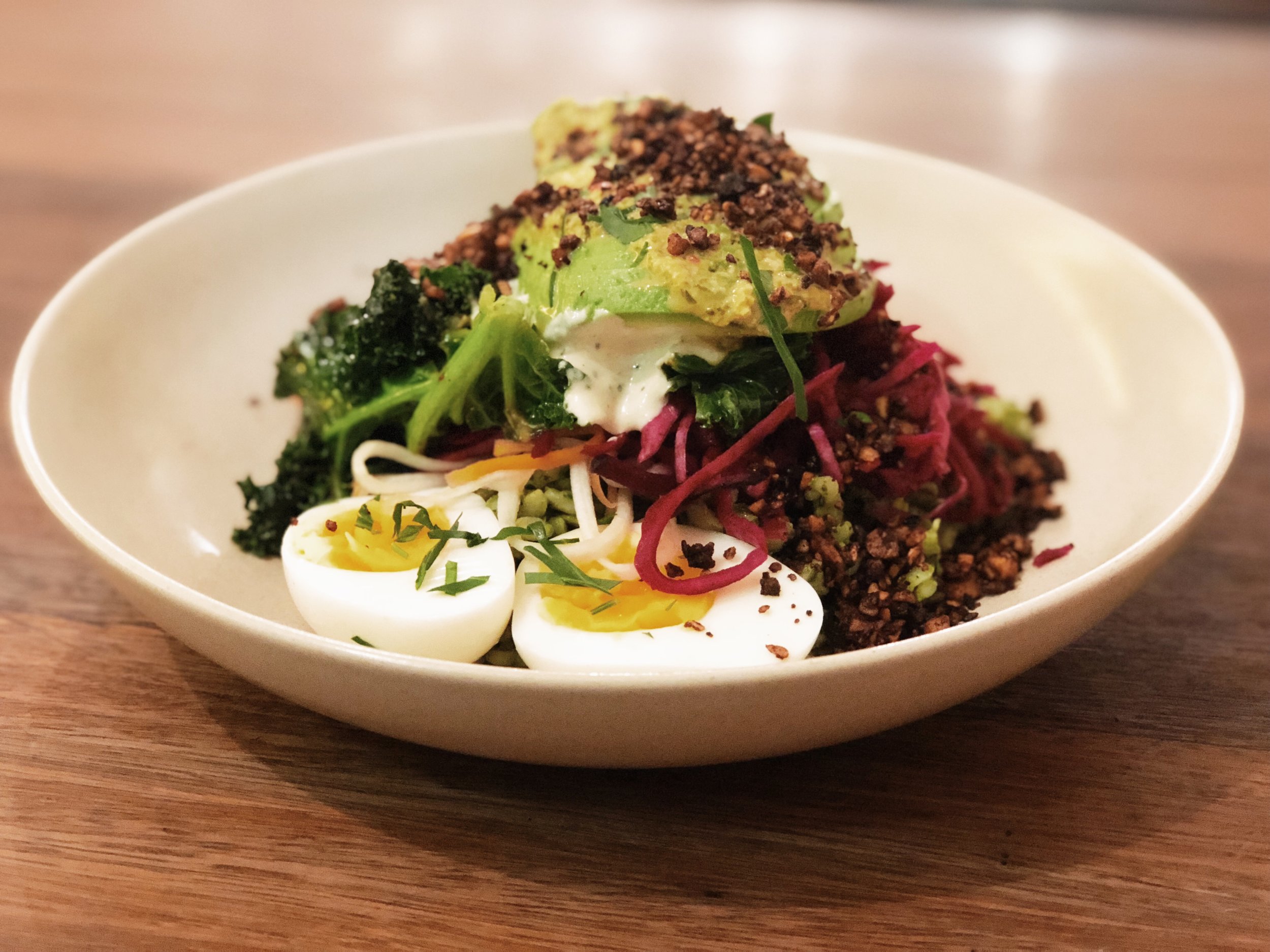 Overhead view of a bowl of nutritious food featuring avocado, greens, noodles, beetroot, and hard-boiled eggs, served on a wooden surface.