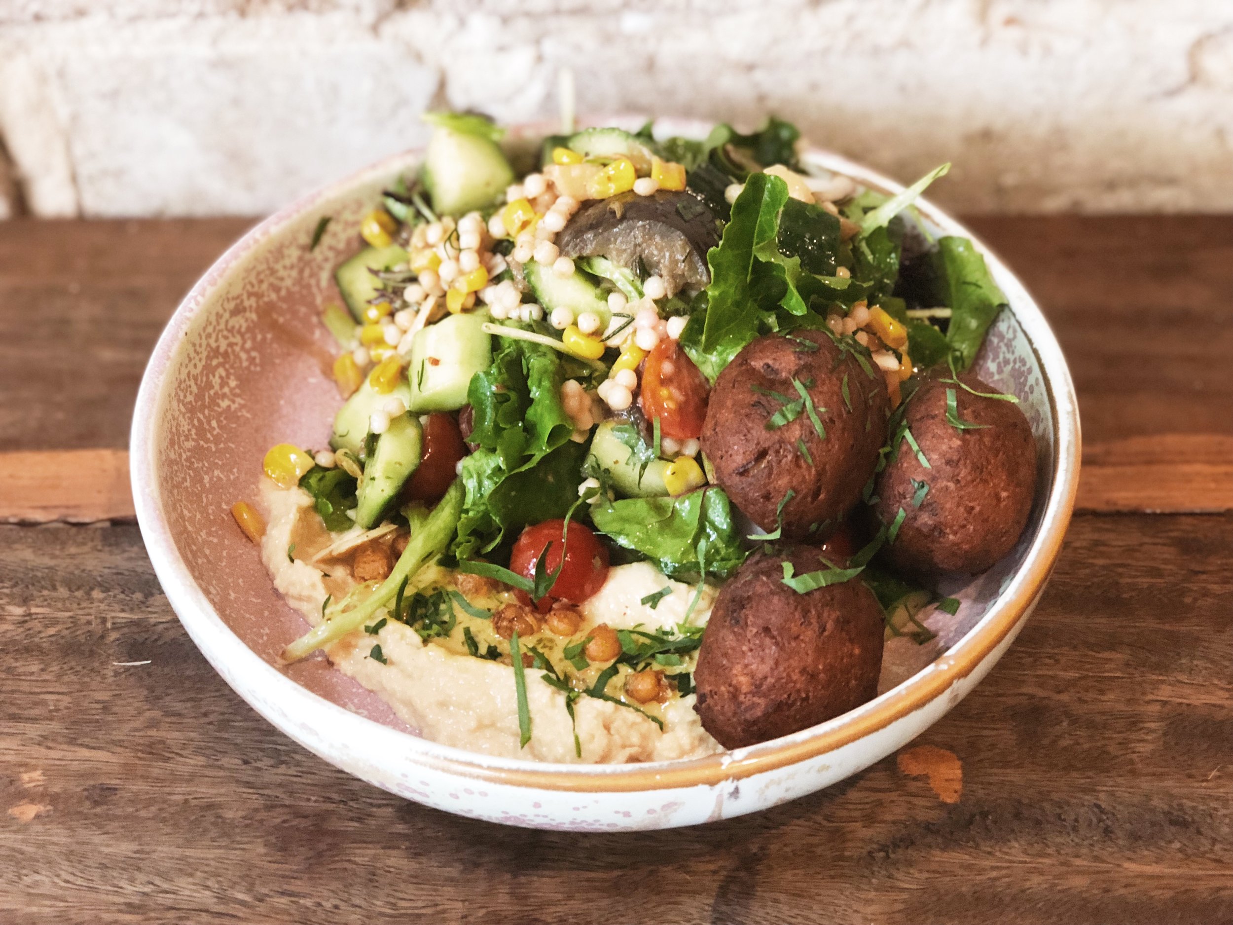 Overhead shot of a bowl filled with salad, couscous, hummus, and falafel on a rustic wooden table. Fresh, colourful, and appetizing food presentation.