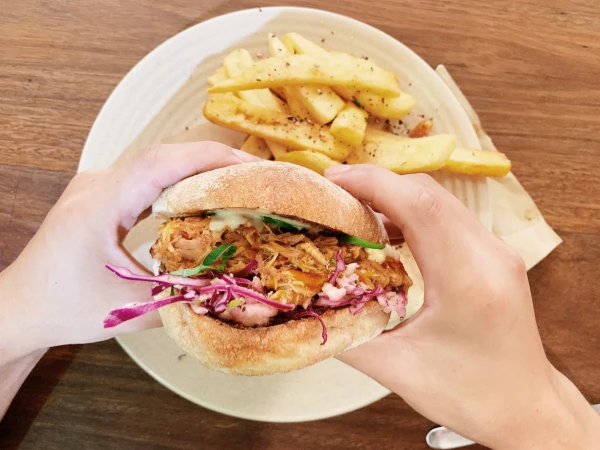Close-up of a pulled pork sandwich with red cabbage held in two hands, served on a plate with seasoned French fries.
