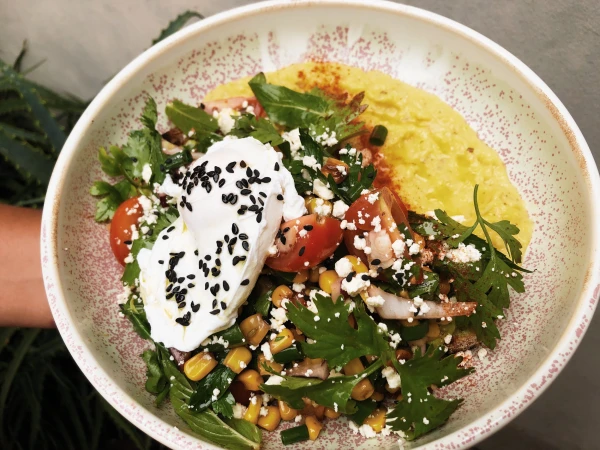 Overhead view of a vibrant salad in a speckled ceramic bowl, featuring a poached egg, corn, tomatoes, greens, and yellow sauce.