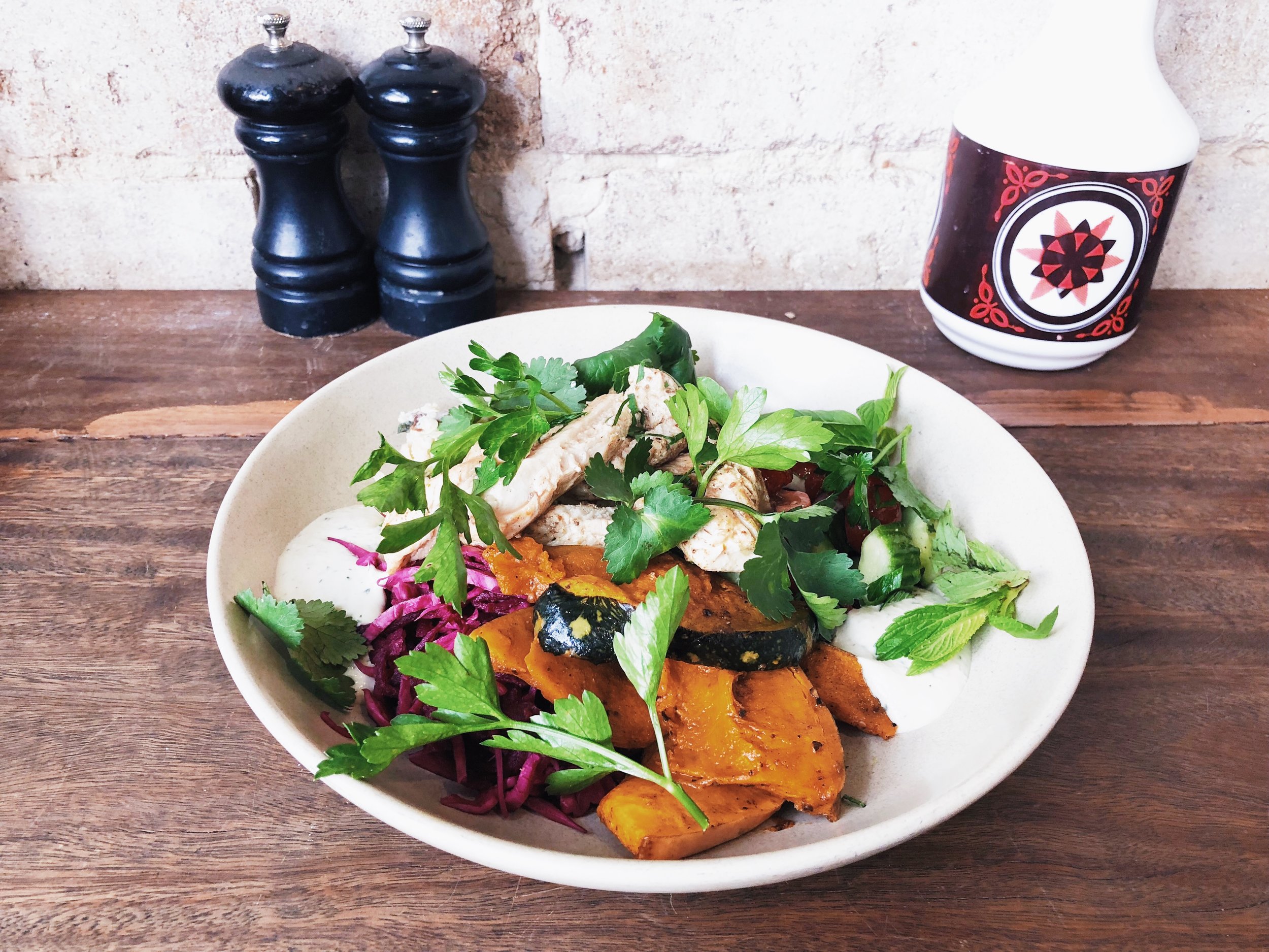 Overhead shot of a white bowl filled with colorful food, including leafy greens, sliced chicken, diced vegetables, and red cabbage on a wooden table.