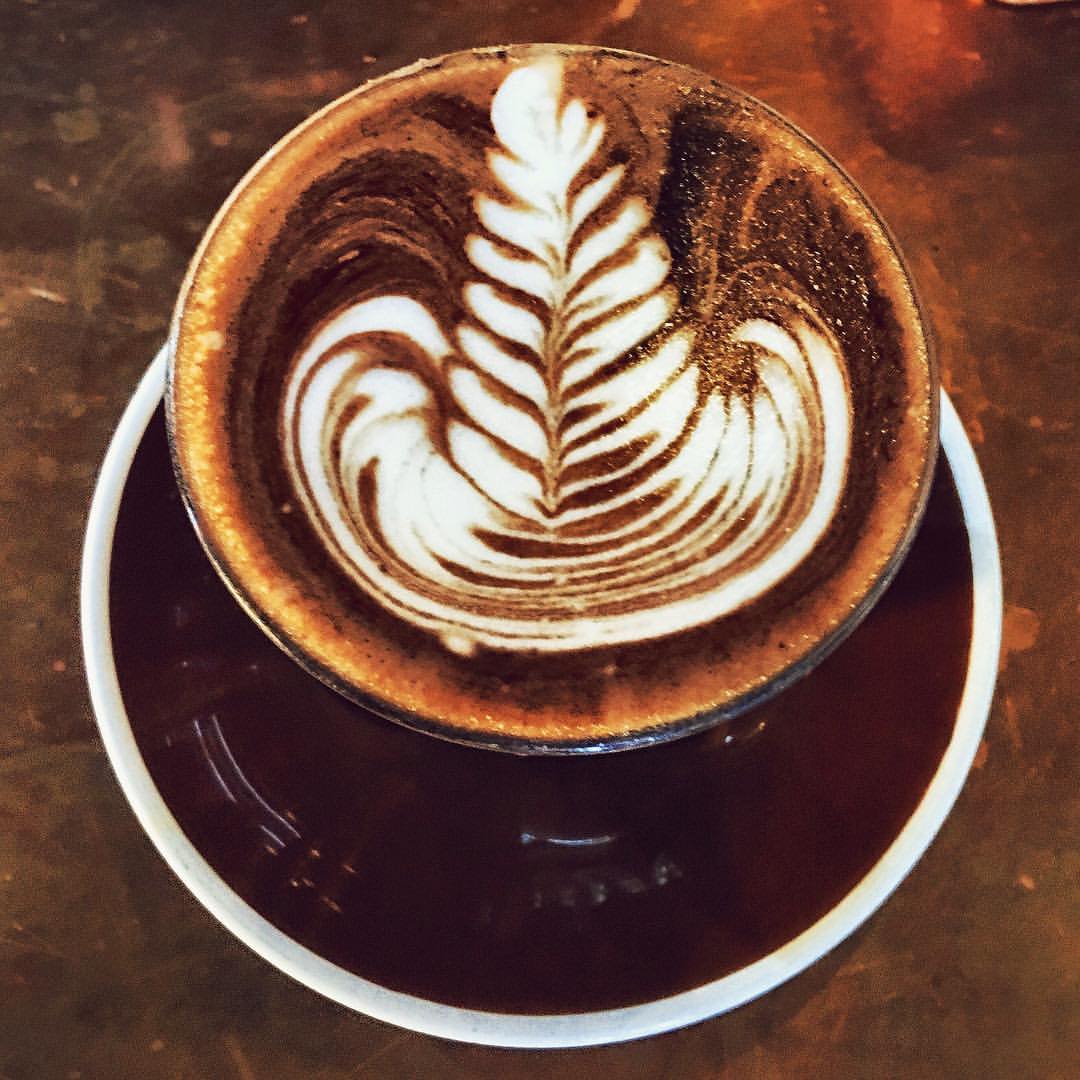 Overhead shot of a latte with leaf latte art, served on a dark brown saucer, sitting on a brown textured surface.