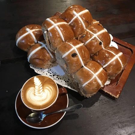 Overhead shot of hot cross buns on a wooden board next to a cappuccino with latte art on a dark surface, food and beverage.