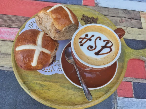 Overhead shot of a cappuccino with latte art, coffee beans, and two hot cross buns on a round wooden tray set on a wood table.