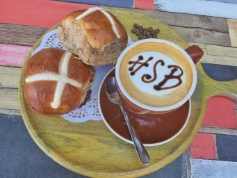Hot cross buns and a cappuccino with latte art on a round wooden serving tray over a multicolored wooden tabletop.