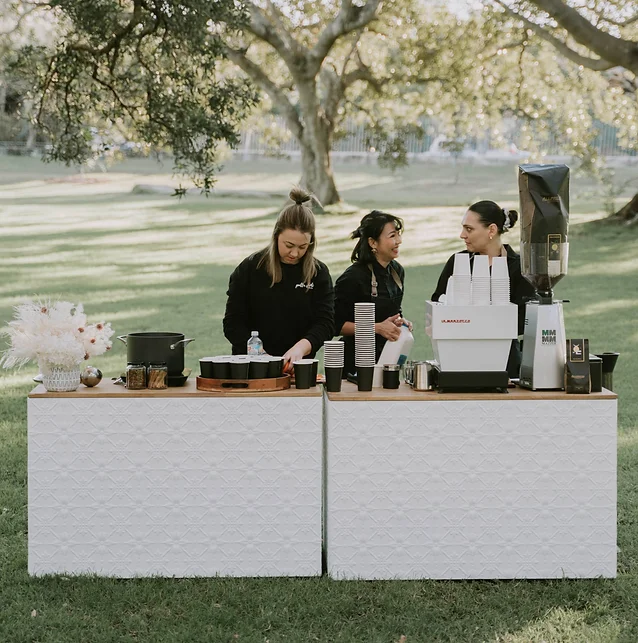 Three women stand behind a serving counter with a coffee machine and supplies in an outdoor setting with a grassy background and large tree.