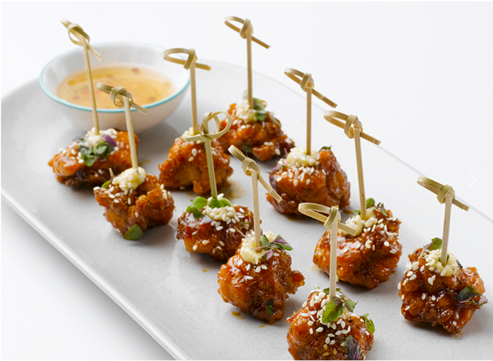 Close-up of savory appetizers: glazed chicken bites on skewers, garnished with herbs and sesame seeds, served on a white rectangular plate.