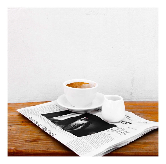 Close-up of a white cup of coffee and a small pitcher on a newspaper atop a wood table with a white wall background.